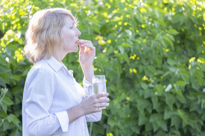 A woman takes a pill washed down with water from a glass in the early morning against a background of greenery