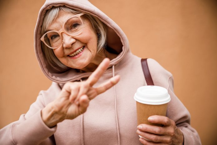 Jolly aged female in hood and glasses is demonstrating peace gesture while drinking coffee