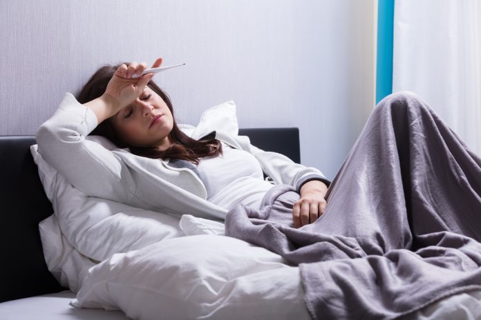 Sick Young Woman Lying On Bed Holding Thermometer In Bedroom