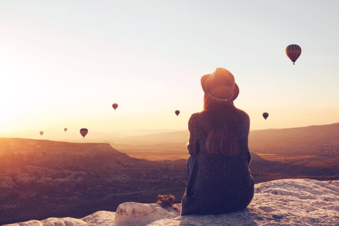 Blick von hinten auf ein Mädchen mit Hut, das auf einem Hügel sitzt und sich Luftballons in Kappadokien in der Türkei ansieht.