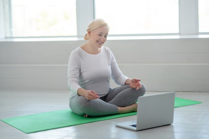 Yoga practice. Mature blonde female sitting in lotus pose, having online yoga lesson, smiling
