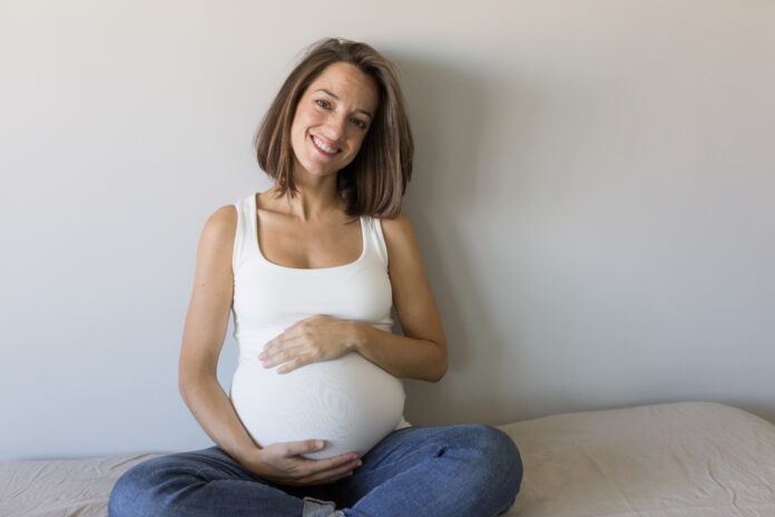 Attraktive schwangere Frau sitzt im Bett, hält ihren Bauch und lächelt. Letzte Monate der Schwangerschaft.Lifestyle drinnen.