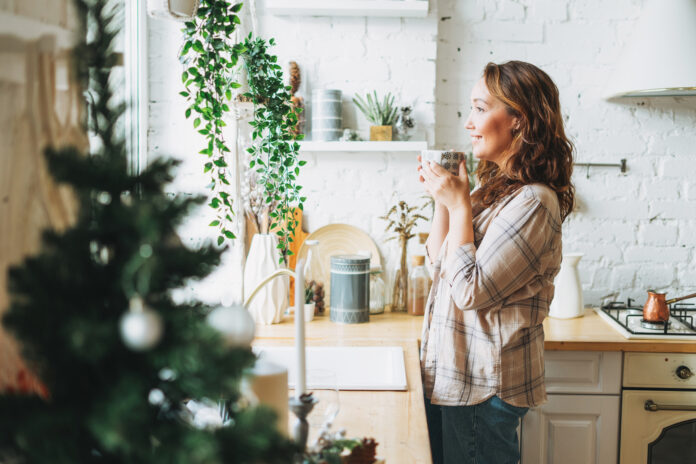 Atractiva mujer sonriente con el pelo rizado en la camisa a cuadros con la taza de té cerca de la ventana en la cocina brillante en casa. Tiempo de Navidad