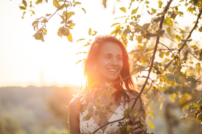 Schöne reife Frau in weißen ärmellosen Kleid stehen entlang Apfelbaum während Sonnenuntergang