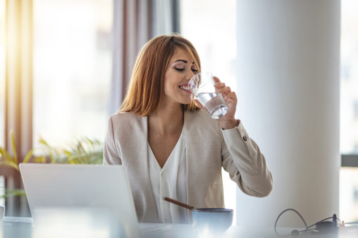 Businesswoman drinking a glass of water at her desk in the office. Young woman working in her offfice. Woman drinking water from glass in the office in the morning with sunlight