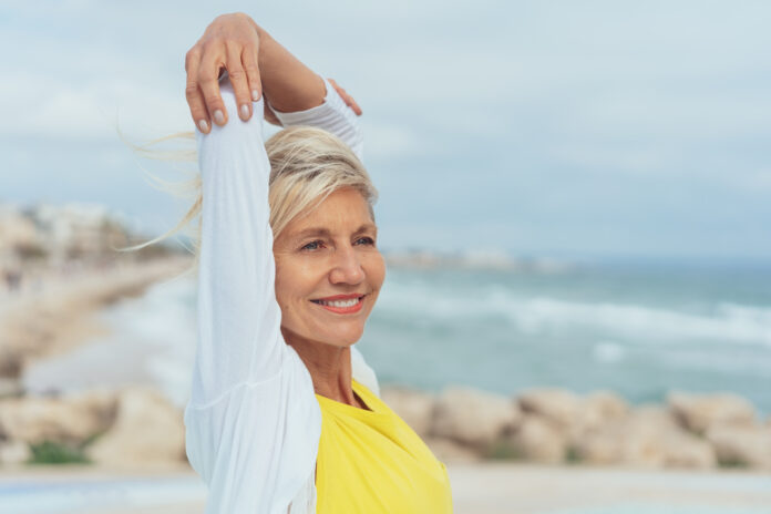 Happy attractive woman at the seaside stretching her arms over her head with a smile as she looks to the side