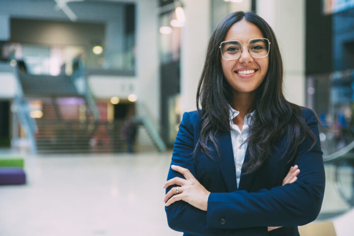 Happy successful businesswoman posing in office hall. Young Latin woman wearing formal suit and glasses, standing for camera with arms folded, smiling. Business portrait concept