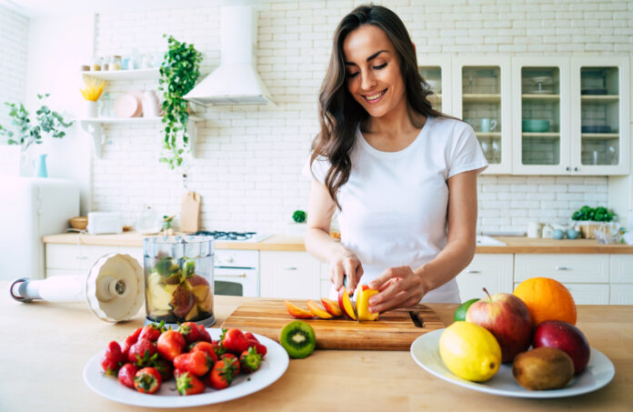 Gesunde Ernährung Lebensstil Konzept Porträt der schönen jungen Frau bereitet Getränk mit Bananen, Erdbeeren und Kiwi zu Hause in der Küche.