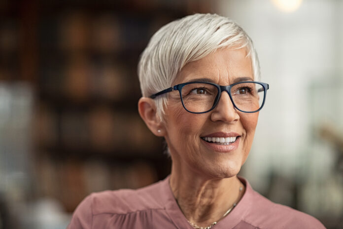 Porträt einer lächelnden reifen Frau mit Brille und kurzen grauen Haaren in einer Bibliothek, die wegschaut. Ältere Bibliothekarin steht im Lesesaal und denkt nach. Alte schöne Dozentin, die in der Bibliothek nachdenkt. Zukunft und Vision Konzept.
