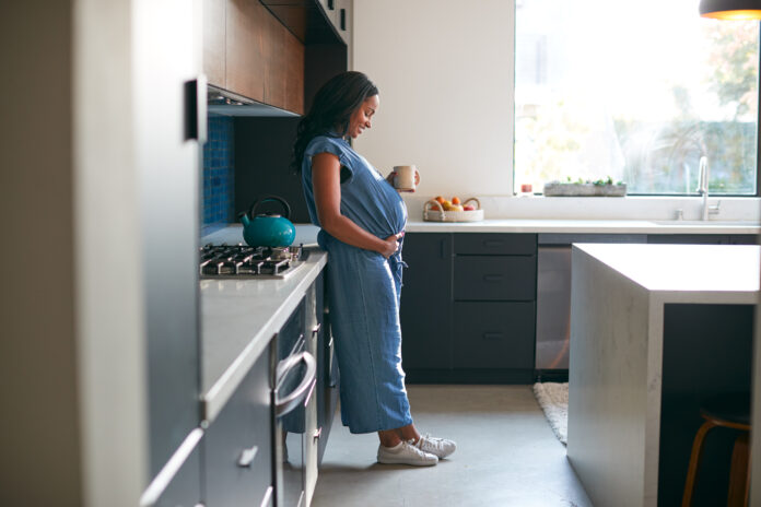 Pregnant African American Woman Standing In Kitchen At Home With Hot Drink
