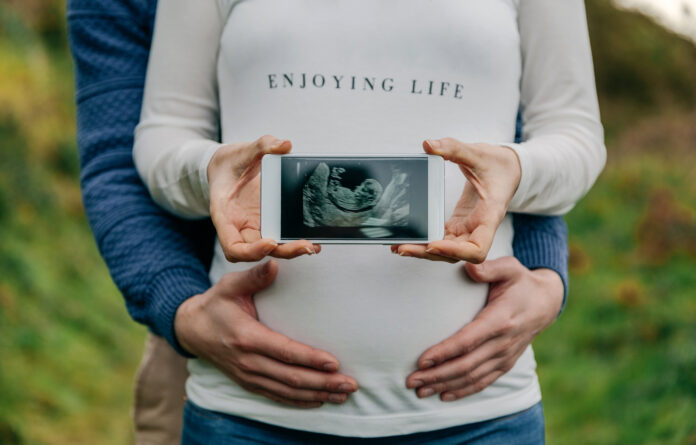 Pregnant woman showing ultrasound of her baby on the mobile embraced by her partner