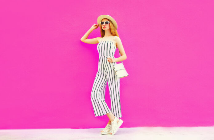 stylish woman model in summer round straw hat, white striped jumpsuit posing on colorful pink wall background