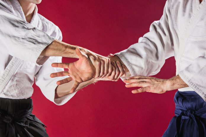 Two men fighting at Aikido training in martial arts school. Healthy lifestyle and sports concept. Men in white kimono on red background. Male hands closeup on red studio background