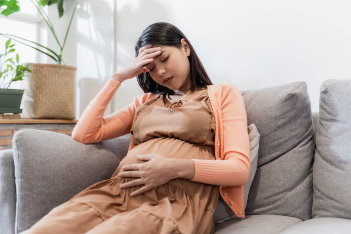 Junge schwangere Asiatin hat beim Sitzen auf dem Sofa unter Kopfschmerzen gelitten. Schwangerschaftssymptome