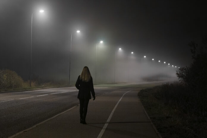 Junge Frau allein langsam zu Fuß unter weißen Straßenlaternen in der Nacht. Dunkle Zeit. Friedliche Atmosphäre im Nebel. Neblige Luft. Rückansicht.