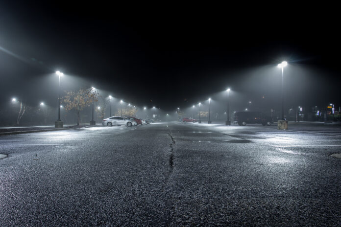 A Lonely Parking Lot on a Cold, Rainy Night - with a Small Collection of Parked Cars in the Background and a Thick Mist Gathering Under the Lights