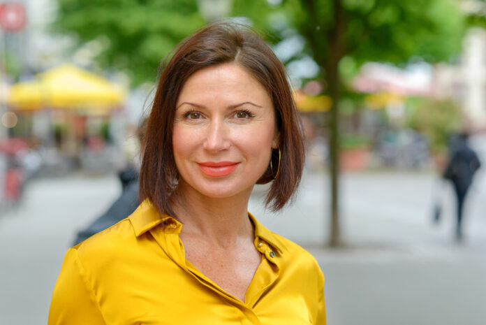 Attraktive reife Frau in buntem gelben Hemd, die einen Tag in der Stadt genießt, steht auf einem ruhigen städtischen Platz und lächelt in die Kamera