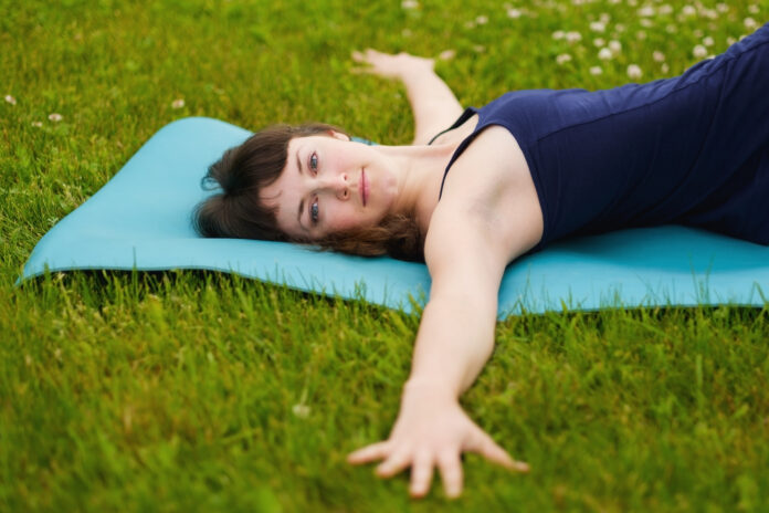 Beautiful caucaisan young woman working out outdoors on blue mat. Girl lying in Reclining Spinal Twist, Jathara Parivartanasana on green grass in park