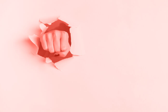 Female fist punching through trendy coral color paper background. War, struggle, conflict, feminist concept. Banner with copy space.