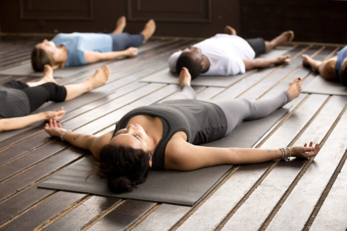Gruppe junger afroamerikanischer und kaukasischer sportlicher Menschen, die eine Yogastunde in toter Körperhaltung liegend praktizieren, Savasana-Übung, Training, Ausruhen nach der Praxis, Innenaufnahme, Studio