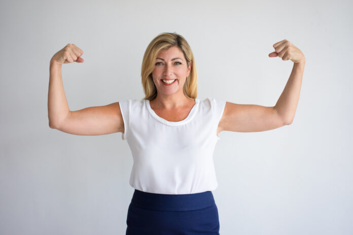 Happy proud Caucasian woman in casual wear flexing biceps. Joyful female consumer advertising food supplement or vitamins for mature women. Healthy and energy concept