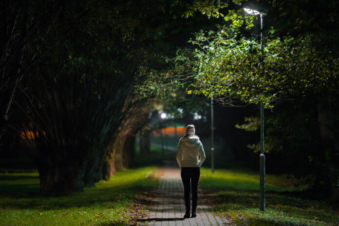 One young alone woman in white jacket walking on sidewalk through alley of trees under lamp light in autumn night. Spending time alone in nature. Peaceful atmosphere. Back view.