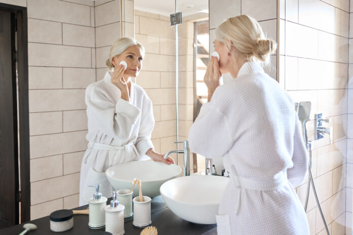 Senior mature older caucasian woman cleansing face with cotton pad removing makeup looking at mirror wearing bathrobe. Everyday routine, anti wrinkle prevention skin care products concept.