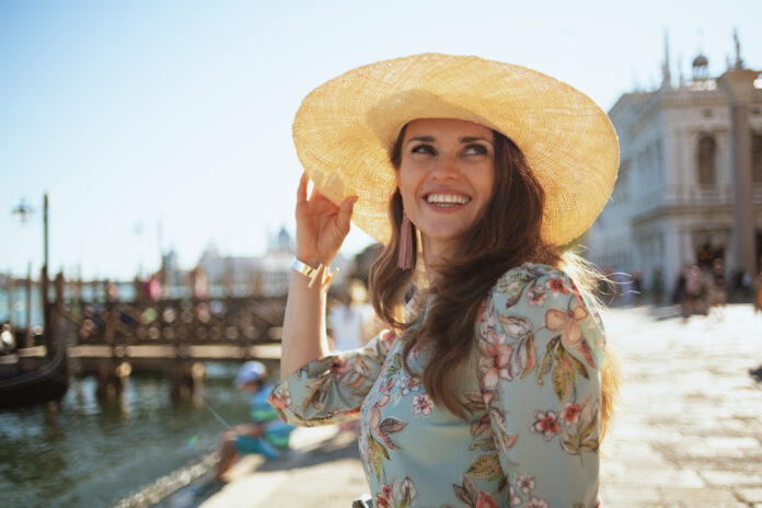 smiling modern middle aged traveller woman in floral dress with hat having excursion on embankment in Venice, Italy.