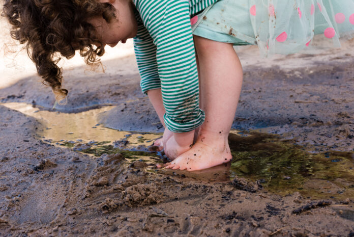 Toddler girl with curly hair and pink and green polkadot skirt playing in sand and water (cropped)