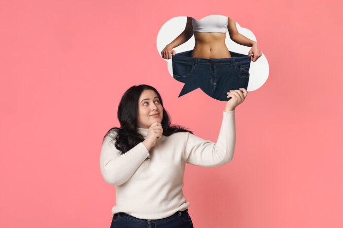Weight Loss Concept. Overweight woman dreaming about slim fit body, holding and looking at her dream sporty figure in speech bubble, pink background