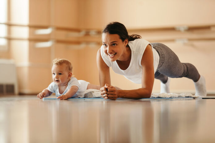 Athletic mother exercising in plank position while being with her baby on sports training. Copy space.