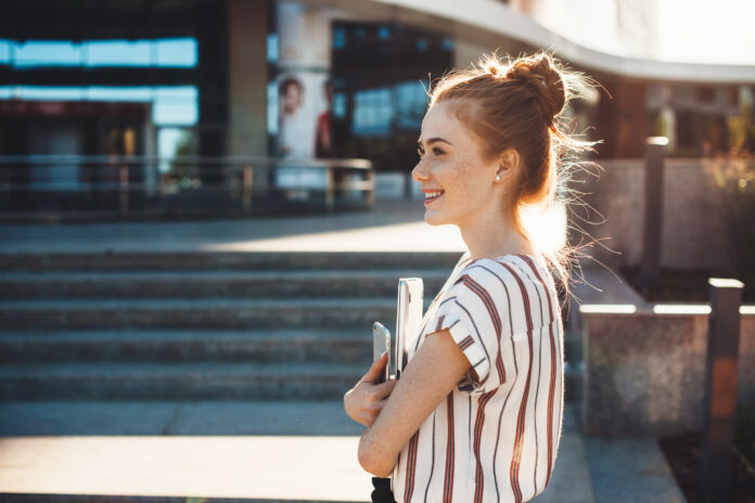 Cheerful caucasian girl with red hair and freckles is holding a tablet and looking away staying in front of a building