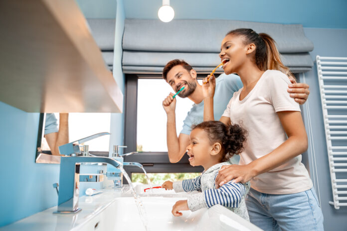 Concept of mixed race family. Low angle view of cheerful young adult man hugging happy woman and brushing teeth at morning. Father and mother standing together with african daughter in bathroom