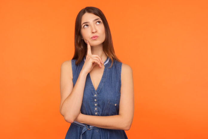 Pensive brunette woman in denim dress looking up with thoughtful doubtful expression, pondering serious difficult idea, imagination and vision in mind. indoor studio shot isolated on orange background