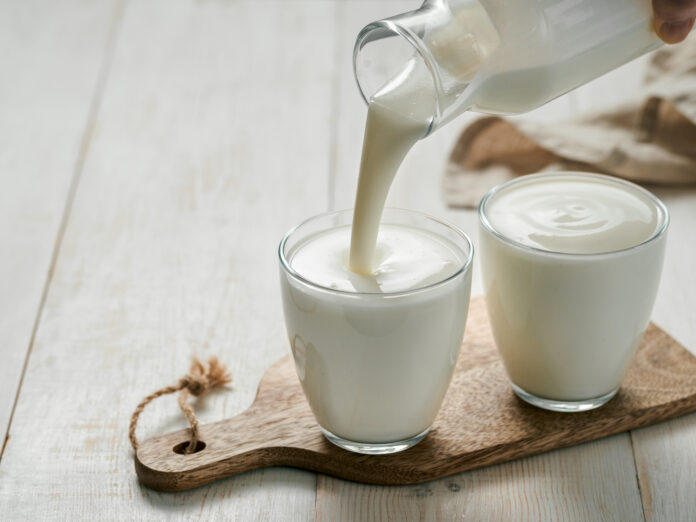 Pouring hausgemachten Kefir, Buttermilch oder Joghurt mit Probiotika. Joghurt fließt aus Glasflasche auf weißem Holz Hintergrund. Probiotisches kaltes fermentiertes Milchgetränk. Trendiges Essen und Trinken. Kopierbereich links