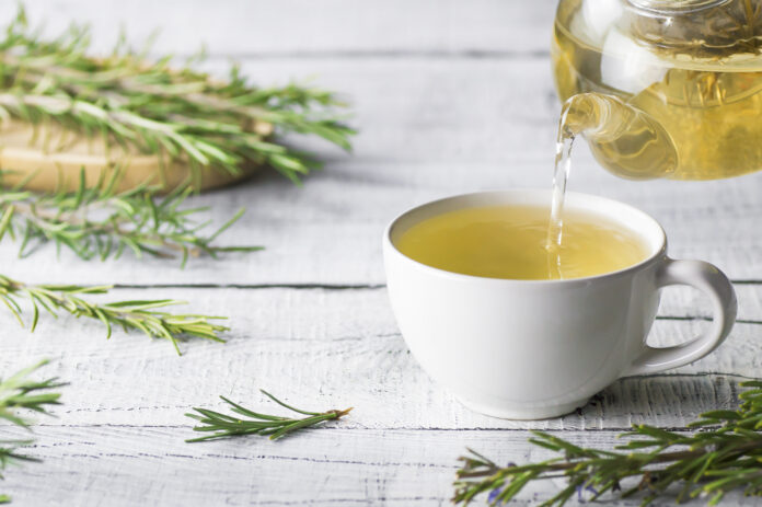 White cup of healthy rosemary tea pouring from teapot with fresh rosemary bunch on white wooden rustic background, winter herbal hot drink concept, salvia rosmarinus