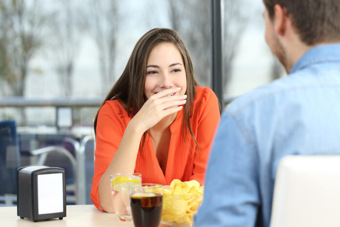 Woman covering her mouth to hide smile or bad breath during a date in a coffee shop with a window in the background