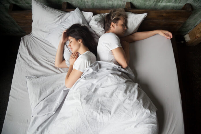 Ung tjej och kille, par som sover under filt med ryggen mot varandra i sängen i sovrummet i hemmet, uppifrån. Tidig morgon, lat söndag helg eller dagslur. Människor som kopplar av och vilar hemma.