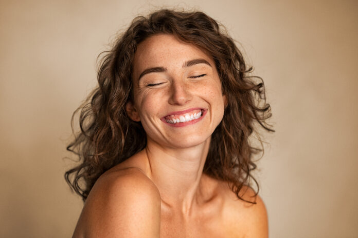 Close up of carefree young woman with bare shoulders laughing. Portrait of smiling woman with freckles and closed eyes enjoying beauty treatment. Beautiful girl laughing isolated on background with copy space, skin care and beauty treatment concept.