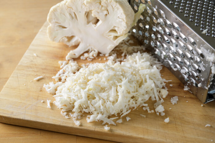 Grating cauliflower with a metal grater on a wooden kitchen board, used as rice replacement for a healthy low carb or ketogenic diet meal, selected focus, narrow depth of field
