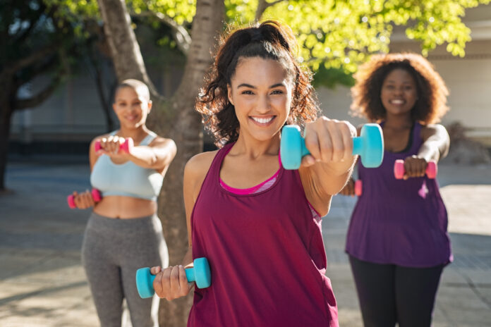 Portrait of young curvy woman with friends exercising with dumbbells. Group of three multiethnic girls working out in park during sunset. Smiling women in sportswear using dumbbell to lose weight.