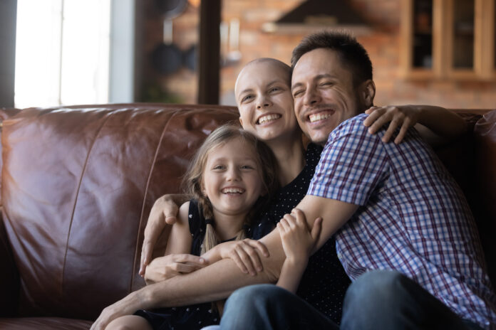 Happy young family with little daughter with sick cancer patient bald mom hug cuddle on couch at home, smiling parents with ill hairless mother relax with small girl child, show love care support