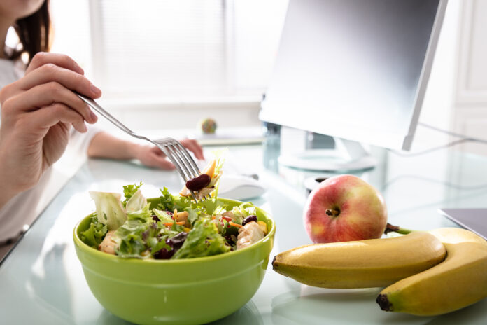 Close-up Of Woman's Hand Holding Fork While Having Healthy Salad On Office Desk