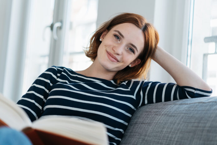 Pretty young woman relaxing on a sofa at home reading a book resting her head on her hand looking at the camera with a sweet sincere smile
