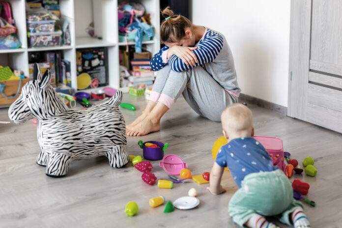 Müde von alltäglichen Haushalt Mutter sitzt auf dem Boden mit den Händen auf dem Gesicht. Kind spielt in unordentlichem Zimmer. Verstreutes Spielzeug und Unordnung. Glückliche Elternschaft.