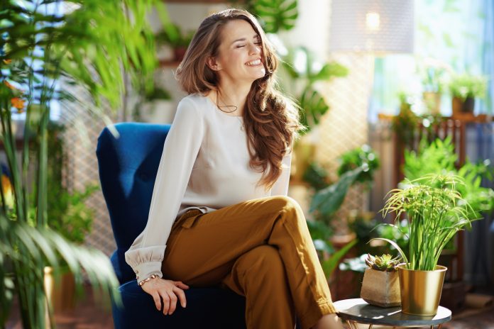 Green Home. smiling elegant woman with long wavy hair at modern home in sunny day in green pants and grey blouse sitting in a blue armchair.