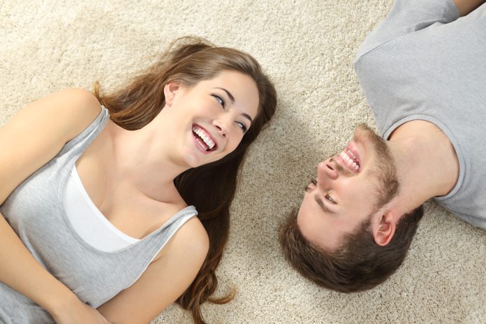 Happy couple laughing looking each other and lying on a carpet at home