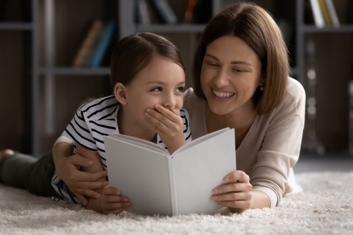 Happy mom teaching little daughter kid to read. Mother and girl relaxing on warm floor at home, reading learning book together, laughing at story, having fun, preparing school homework
