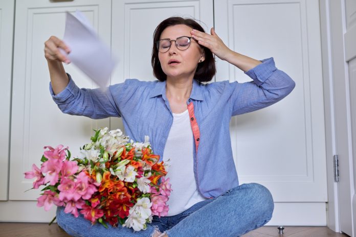 Hot flashes in a woman of mature age, symptoms of menopause, a female sitting at home on the floor blowing her face with papers