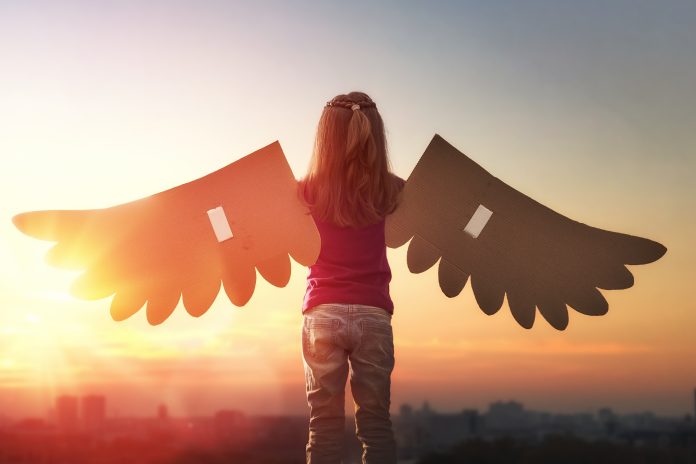 Little girl plays outdoors. Child on the background of sunset sky. Kid with the wings of a bird dreams of flying.
